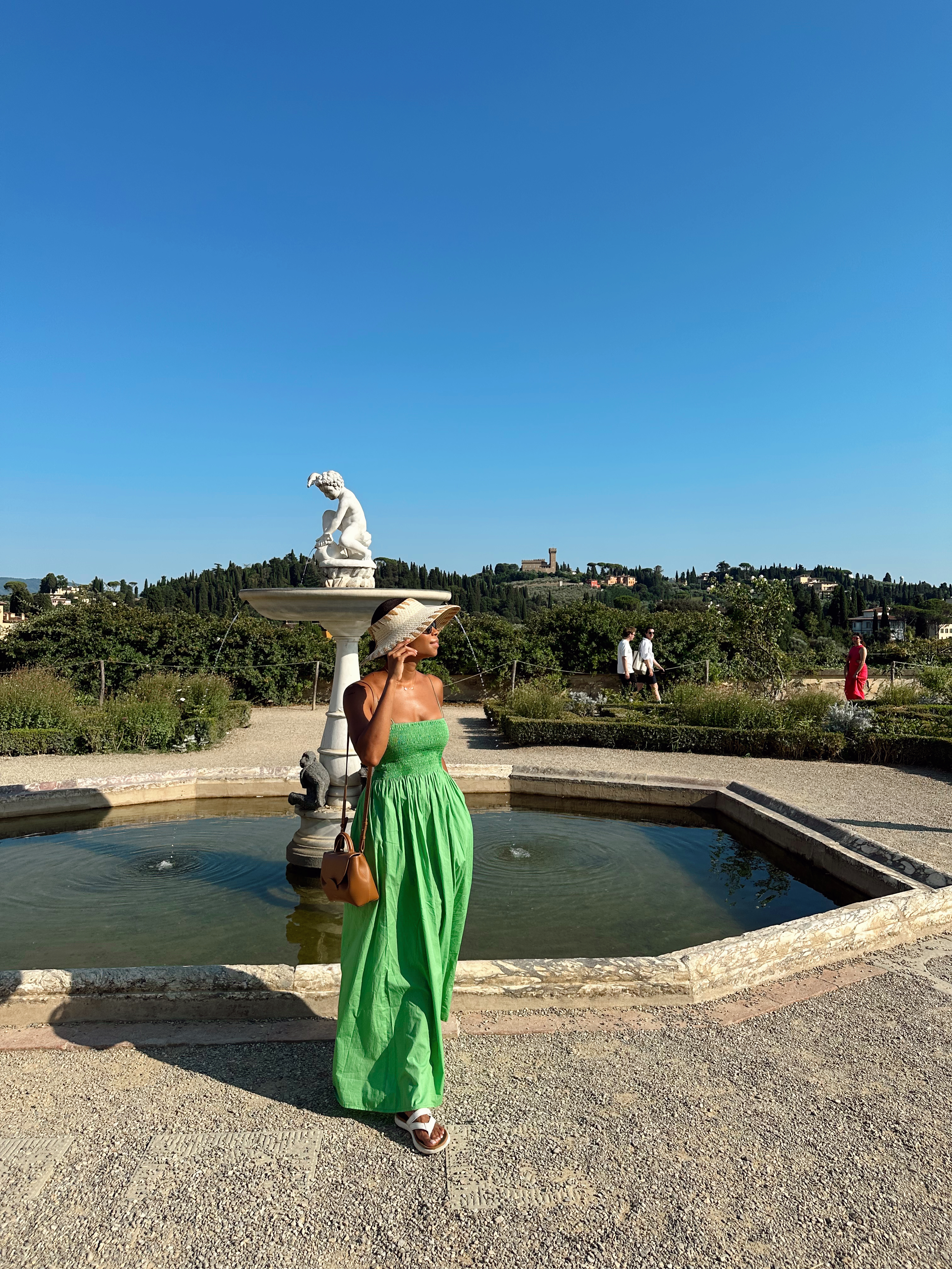 A person in a green maxi dress stands by a fountain in a garden with a clear blue sky in the background.
